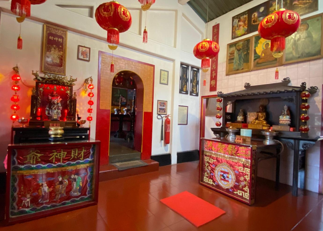 Altar Dewa Rezeki dan patung Buddha menghiasi ruangan dengan lampion merah yang bersinar terang, melambangkan harapan akan kelimpahan dan pencerahan dalam mitologi serta ajaran spiritual Tionghoa.